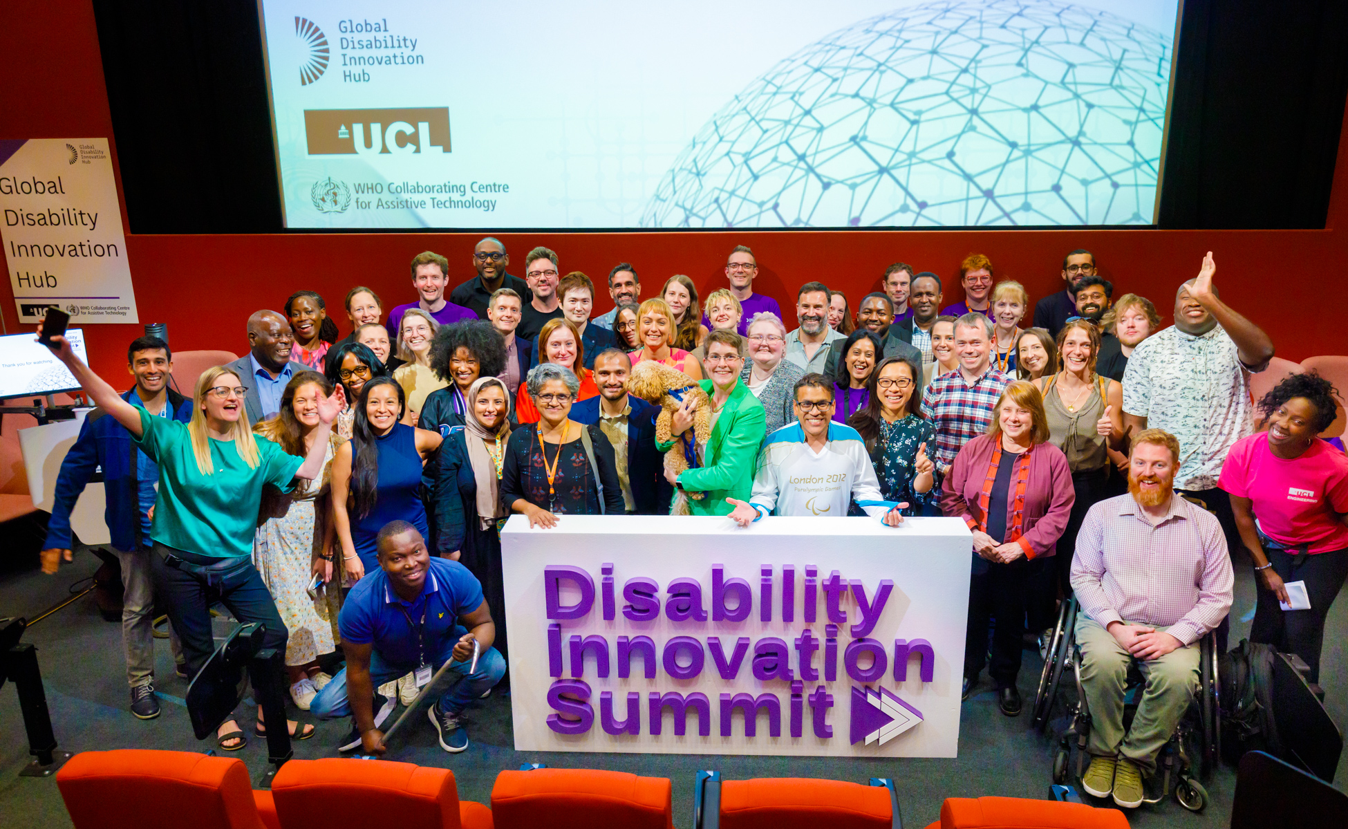 A group of diverse people standing on a stage in front of board saying Disablity Innovation Summit with Global Disability Innovation Hub projected on the screen in the background